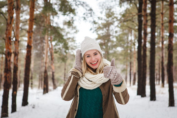 Beautiful young girl outdoors in winter looking at camera smiling and lifting her thumb up, showing cool. Portrait of attractive happy girl outdoors in winter background. Close-up.