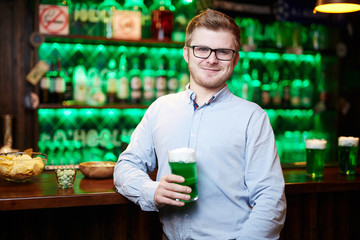 Young irish man in eyeglasses holding glass of green drink with foam and leaning against bar counter