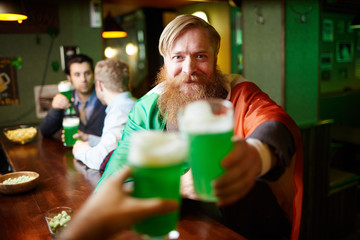 Irish bearded man wrapped into national flag toasting with one of his friends in pub at leisure