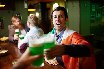 Fototapeta na wymiar Excited young man with irish flag on shoulders cheering up with glass of beer with his friend in bar