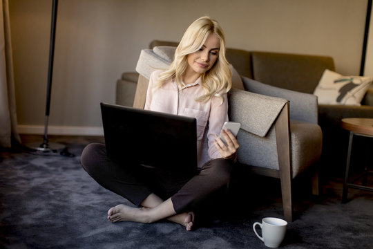 Blonde young woman using laptop and mobile phone while sitting on the floor in the room