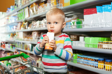 Little boy drinking juice in shopping cart near shelves with baby food at supermarket