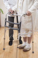 Pielęgniarz trzyma dłoń na ręce staruszki pomagając jej w chodzeniu za pomocą balkonika.