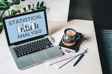 Workplace without people, close-up of laptop with inscription statistical analysis on screen on white table, desk.Nearby is closed paper notebook, pen, newspaper, cup of coffee. Online marketing