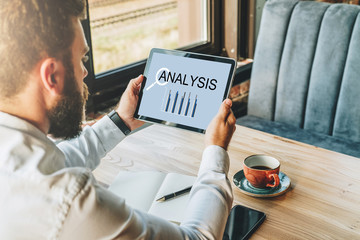 Back view. Young bearded businessman in white shirt is sitting at table, using tablet computer with...