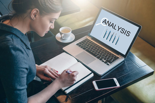 Side view. Young woman sitting at table in cafe in front of laptop with inscription analysis on screen and making notes in notebook, diary. Businesswoman working, student studying.