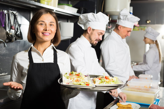 smiling woman waiter collecting dishes from restaurant’s kitchen
