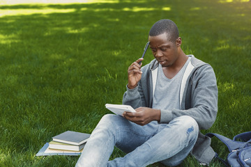 Concentrated african-american studen sitting with notebook on grass