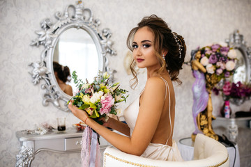 A beautiful bride with a wedding bouquet in her hands sits in a chair, looks into the camera.