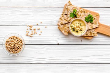 Serve hummus. Bowl with dish near pieces of crispbread on white wooden background top view copy space