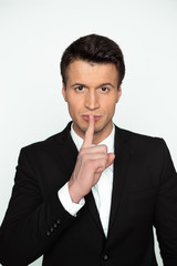 Hush please. A young confident handsome man in a black jacket holds a finger near his lips and looks into the camera.