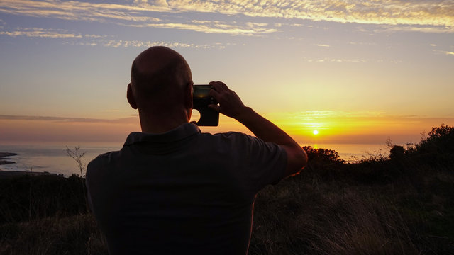 Young man takes a photo to the sunset with a smartphone with the sea in background