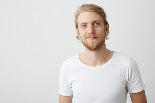 Indoor shot of positive good-looking caucasian blond man with beard and moustache smirking while looking at camera and standing over gray background in white t-shirt. Feelings and emotions concept