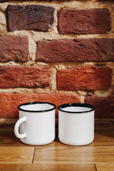 White metal cups stand on a wooden floor, against a red brick wall.