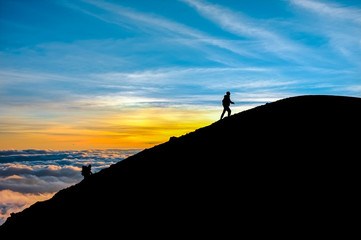 Two people climbing a mountain are almost on the summit. It is sunrise with clouds in the sky. Full of color. 