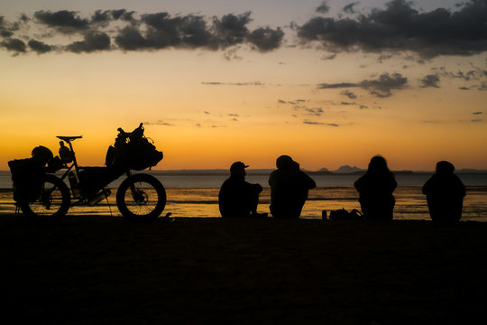 Four people sit on a cliff above the sea in Baja California at sunset next to a loaded touring bicycle with a view of the ocean