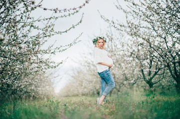 Fototapeta na wymiar Beautiful young cheerful pregnant woman in wreath of flowers on head touching belly while walking in spring tree garden. Beauty People Motherhood concepts. Happy mom in waiting for baby!