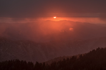 The sun at sunset is partially hidden by dark clouds. It is setting behind a line of mountain ridges. The photo is dark with layers of ridges in the foreground and barren trees of winter