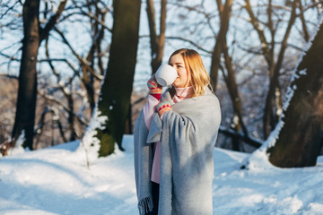 Young caucasian attractive blonde girl in pink sweater and knitted scarf holds white cup of tea in winter park outdoor