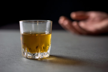 glass of whiskey or cognac or alcohol drink, hand of a drunk man in the background, alcoholism and alcohol abuse concept, defocused, selective focus, close up, gray table, dark background