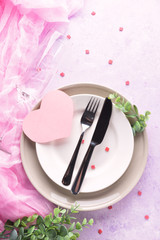 Table setting with a pink paper heart. White and grey plates, champagne glasse, cutlery and decorative textile on ultra violet background