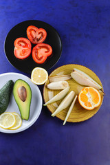 Fototapeta na wymiar Avocado and lemon on a white plate, tomato on a black plate, citrus on an ultraviolet background, chopped banana and pear, avocado pop art, healthy food, vegan, art