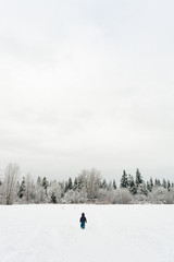 View from behind of a young child walking through a snowy field towards a group of trees in winter