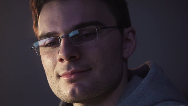 Young unshaved caucasian guy in glasses sitting in front of computer at night start smiling getting an achivement, close up portrait