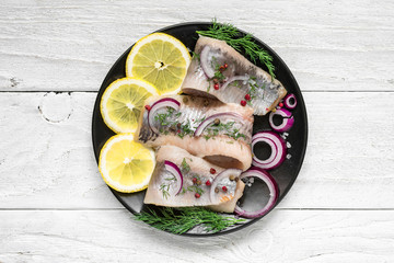 Herring fillet with salt, pepper, herbs, onion and lemon on black plate on white background