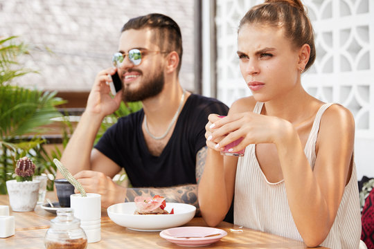 Bored discontent young woman being irritated with boyfriend who always chats on mobile phone, spend free time at cafeteria. Bearded tattooed male model has telephone conversation with someone