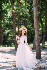 Fototapeta na wymiar Photo of a beautiful young bride in a beautiful wedding dress with a wreath on her head and with a bouquet of flowers in her hands, she stands in the park on a beautiful sunny day
