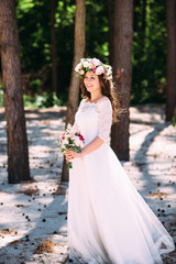 Obraz na płótnie Canvas Photo of a beautiful young bride in a beautiful wedding dress with a wreath on her head and with a bouquet of flowers in her hands, she stands in the park on a beautiful sunny day