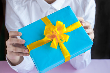 Female hands holding a gift in a blue box with a yellow ribbon on a pink background