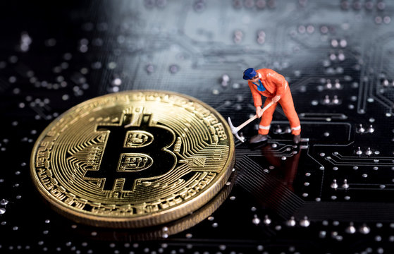 Bitcoin mining miniature worker, small figure holding mattock digging on shiny golden Bitcoin crypto currency coin on electronics cyber look circuit board with soldering