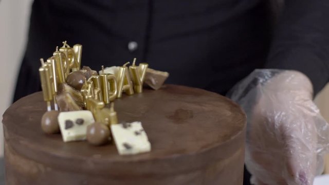 Female confetioner is decorating top of cake for celebration with candies, close up. Lady is turning the baked sweet aroung on the table, putting pieces of white chocolate and bar, and the sign Happy