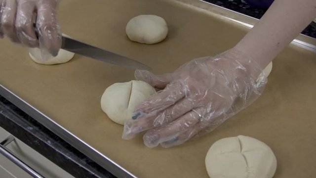 Close up of cuts are being made on the bun by female baker. Five round pieces of dough are on baking pan covered with brown paper and lady in white gloves is using steel knife for decorating the rolls