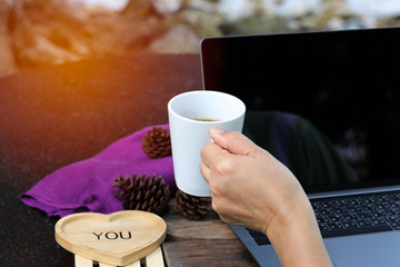 Hand-lifted white hot coffee cup and heart-shaped wooden tray with letter ME on the side laptop has a purple cloth backdrop and pine cones.