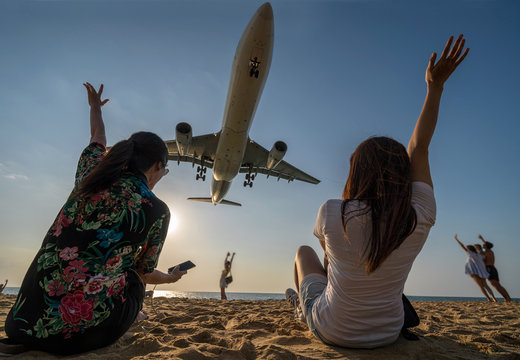 Scene of Two traveler woman showing hand and funning with Airplane landing closely at the sea beach beside the phuket international airport, Thailand