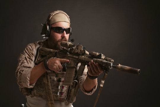 Brutal man in the military desert uniform and body armor stands in a fighting rack and holds his rifle on a black background. The bearded player in the airsoft safety glasses aiming a rifle