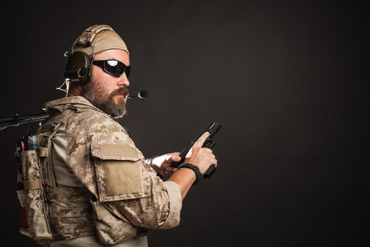 Brutal man in the military desert uniform and body armor stands and holds a gun on a black background in the Studio. The bearded player in the airsoft safety glasses or goggles and active headphones