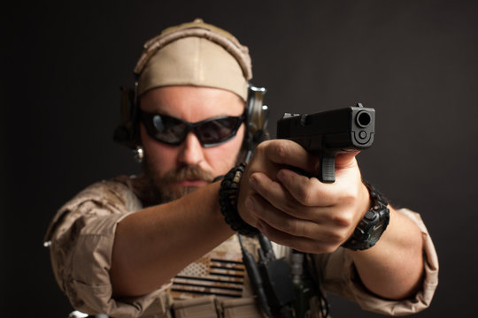 Brutal man in the military desert uniform and body armor standing in a fighting rack and aiming from his gun on a black background. Close-up of the gun barrel directed towards the camera