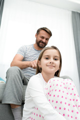 Fototapeta premium Portrait of cheerful bearded father combing hair of teenage daughter in bedroom, with girl looking at camera and smiling