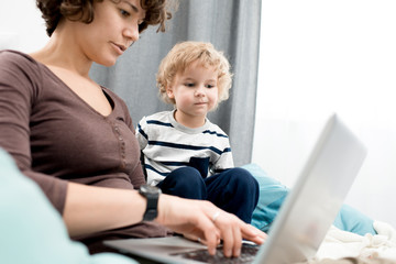 Portrait of young mother using laptop at home, focus on curious little boy looking at screen and...