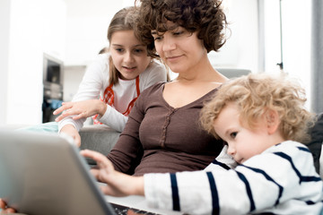 Portrait of young mother using laptop with two children looking at screen while searching internet...