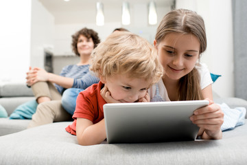 Portrait of brother and sister using digital tablet lying on sofa in living room with parents...