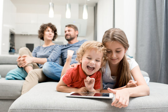 Portrait of brother and sister using digital tablet and laughing while lying on sofa in living room with parents watching TV in background, copy space