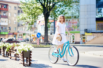 Fototapeta na wymiar Smiling young female in white dress riding blue bike in front of modern city buildings next to flowerbeds on sunny summer day. Beautiful woman turquoise bicycle green trees happiness enjoying ride