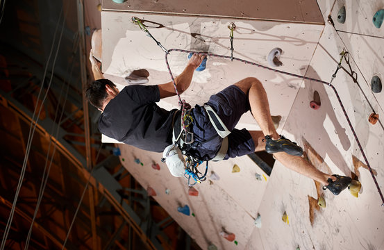 Strong young man pushing himself higher to reach the top of artificial climbing wall in bouldering gym indoors. Motivation healthy sport lifestyle exercise activity hobby effort muscles no limit