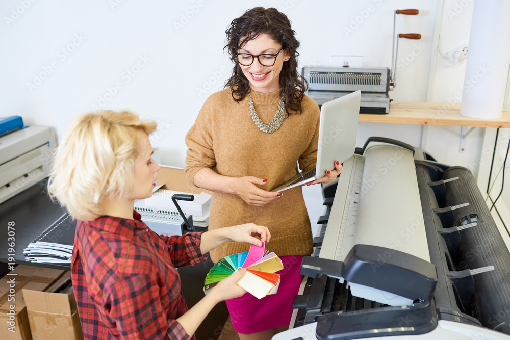 Poster portrait of two graphic designers standing by plotter machine in printing shop, focus on smiling wom
