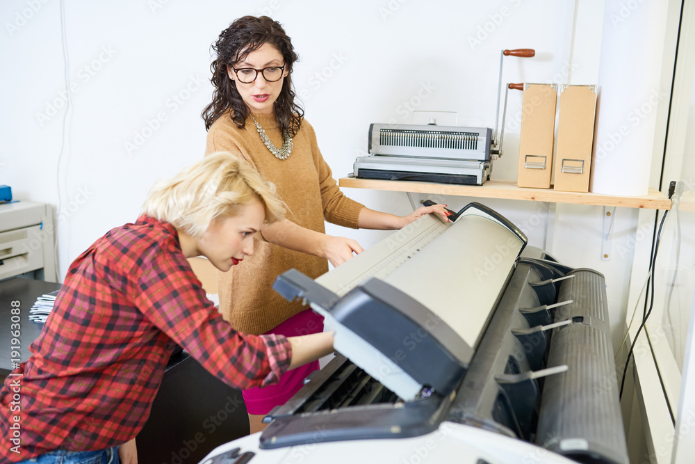 Poster portrait of two women setting up plotter machine in printing shop, focus on manager giving instructi